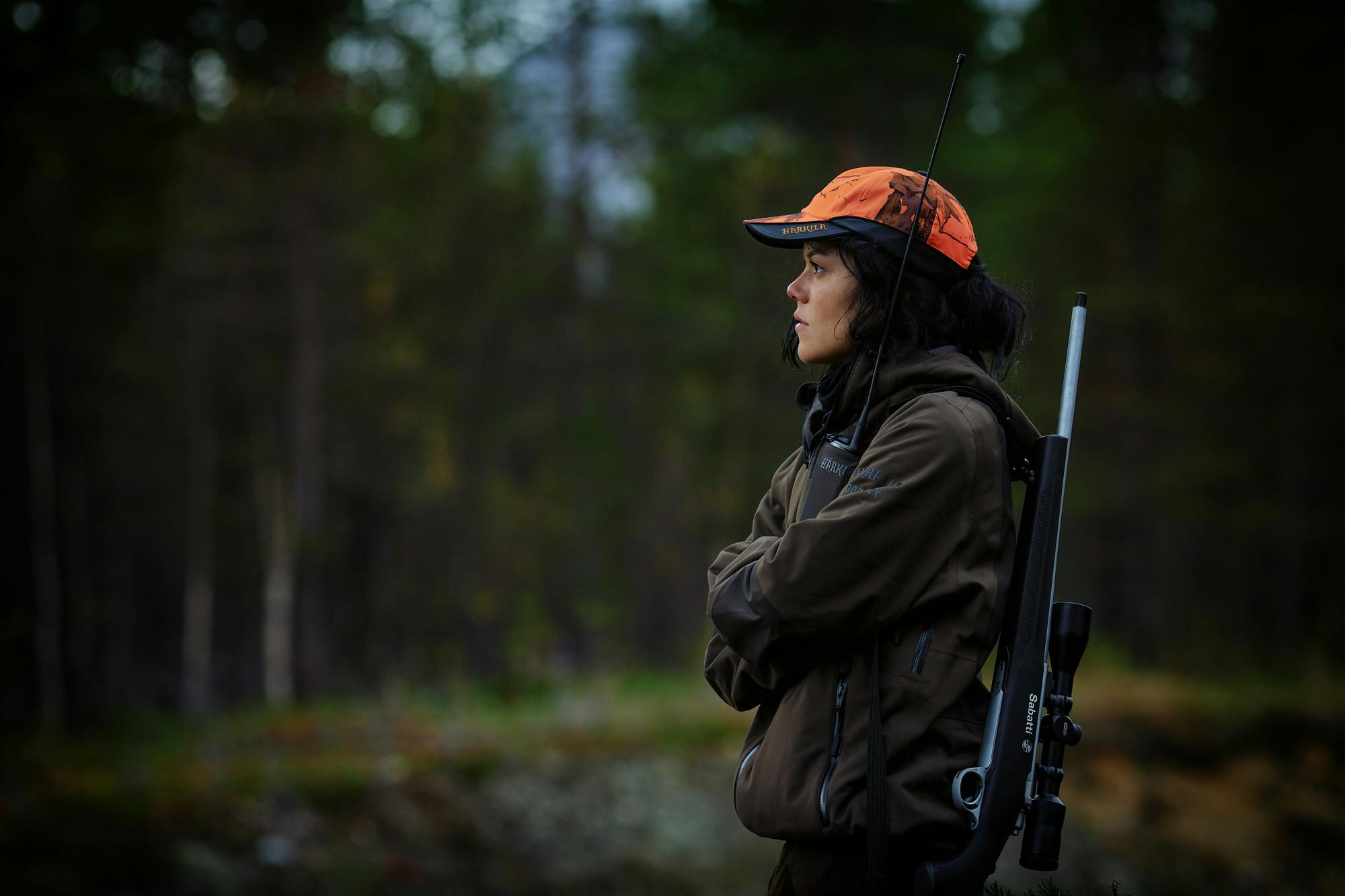 A hunter in a blaze-orange cap with a scoped rifle on her shoulder, standing at the edge of a dark spruce forest.