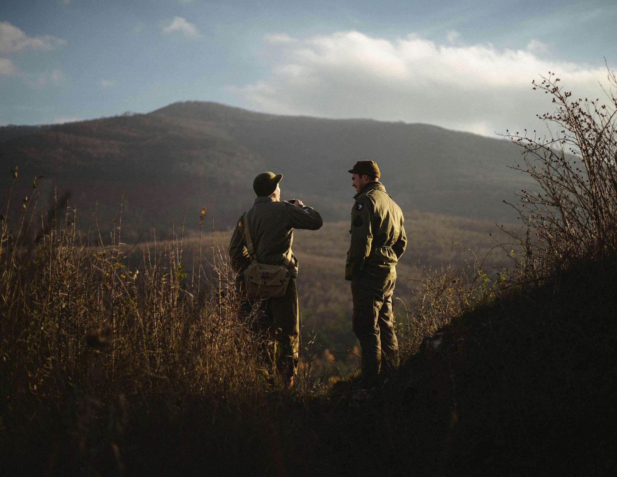Two hunters on a ridge at dusk, one pointing across the valley toward a far hillside.
