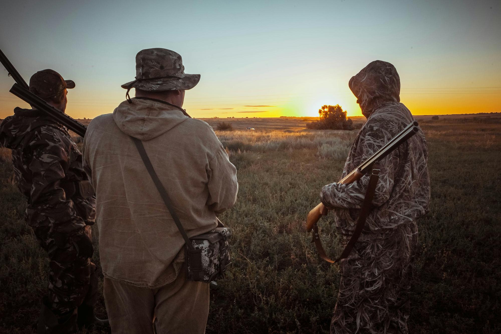 Three hunters with shotguns standing in a field at sunrise.